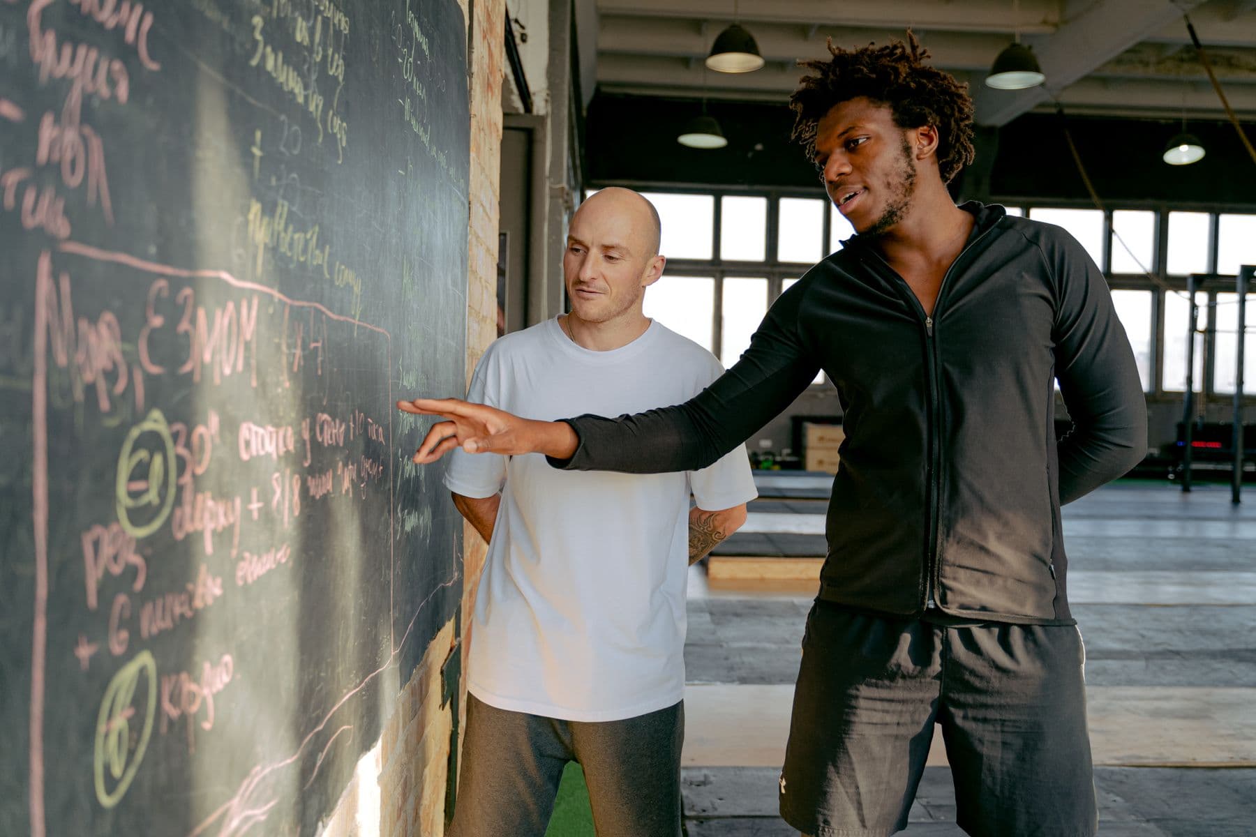 A coach walking a client through the session written up on a gym blackboard
